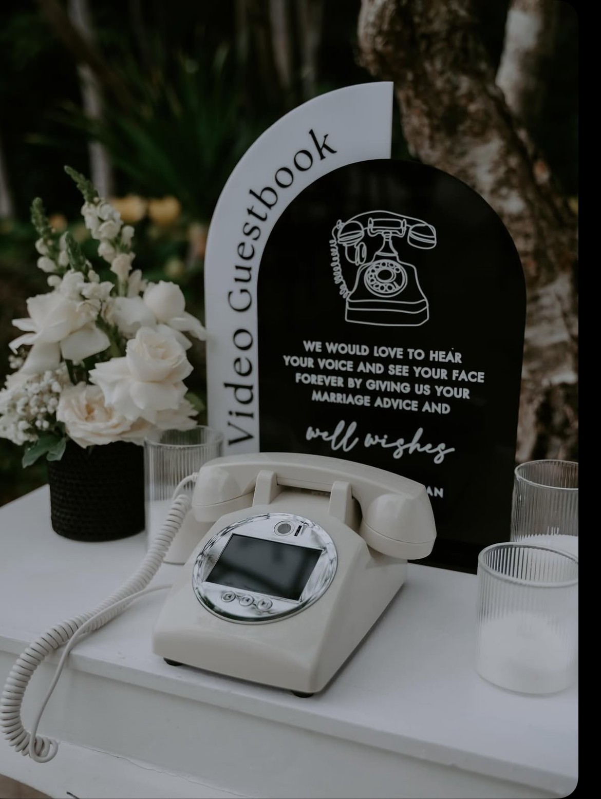 A cream rotary-style video guestbook phone on a table beside white florals, with an arched sign reading 'Video Guestbook'.